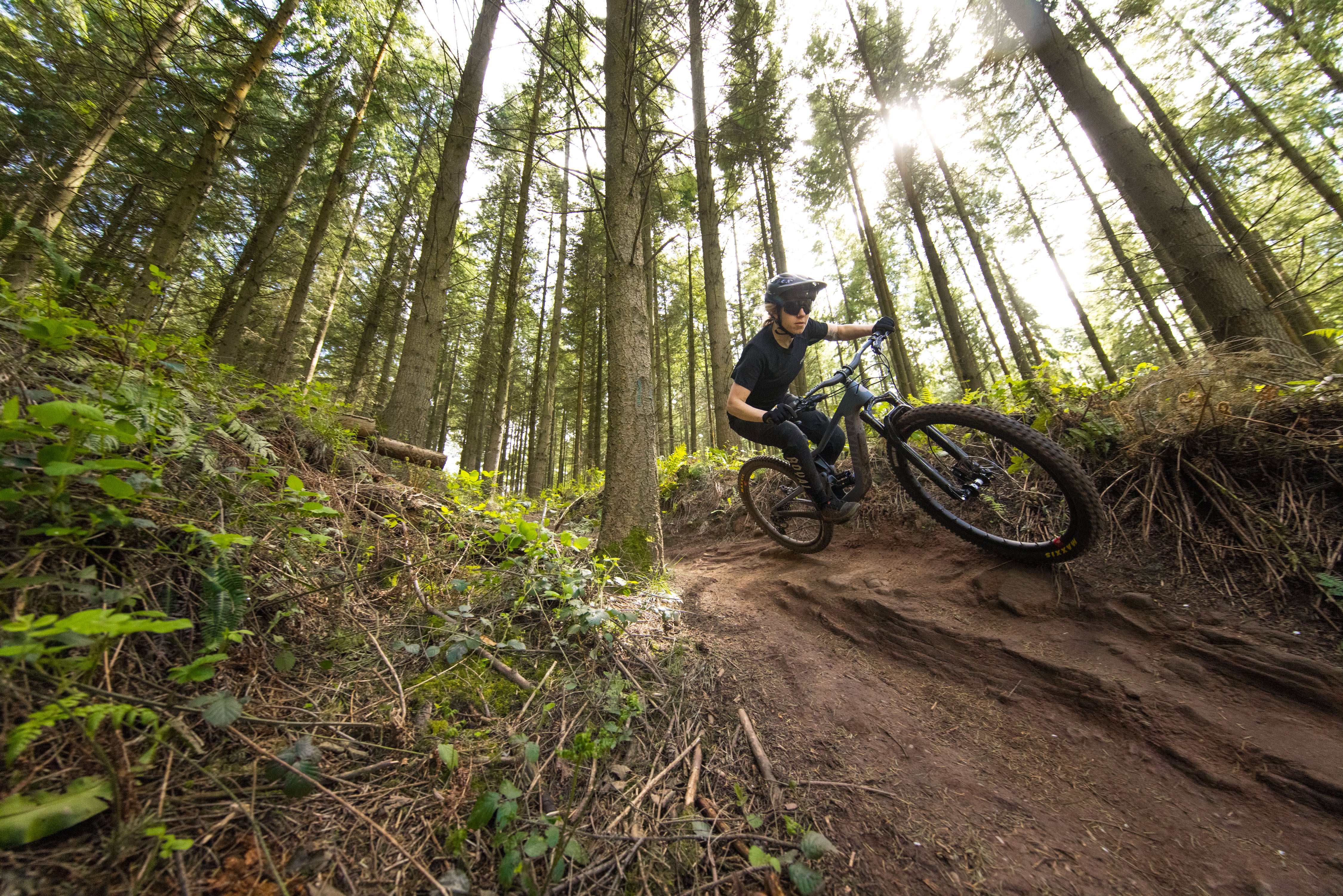 An action shot of a woman navigating a scenic trail on a Juliana Roubion mountain bike, surrounded by tall grass with towering mountains in the distance, showcasing the bike’s capability and performance on diverse terrain, available at Contender Bicycles.