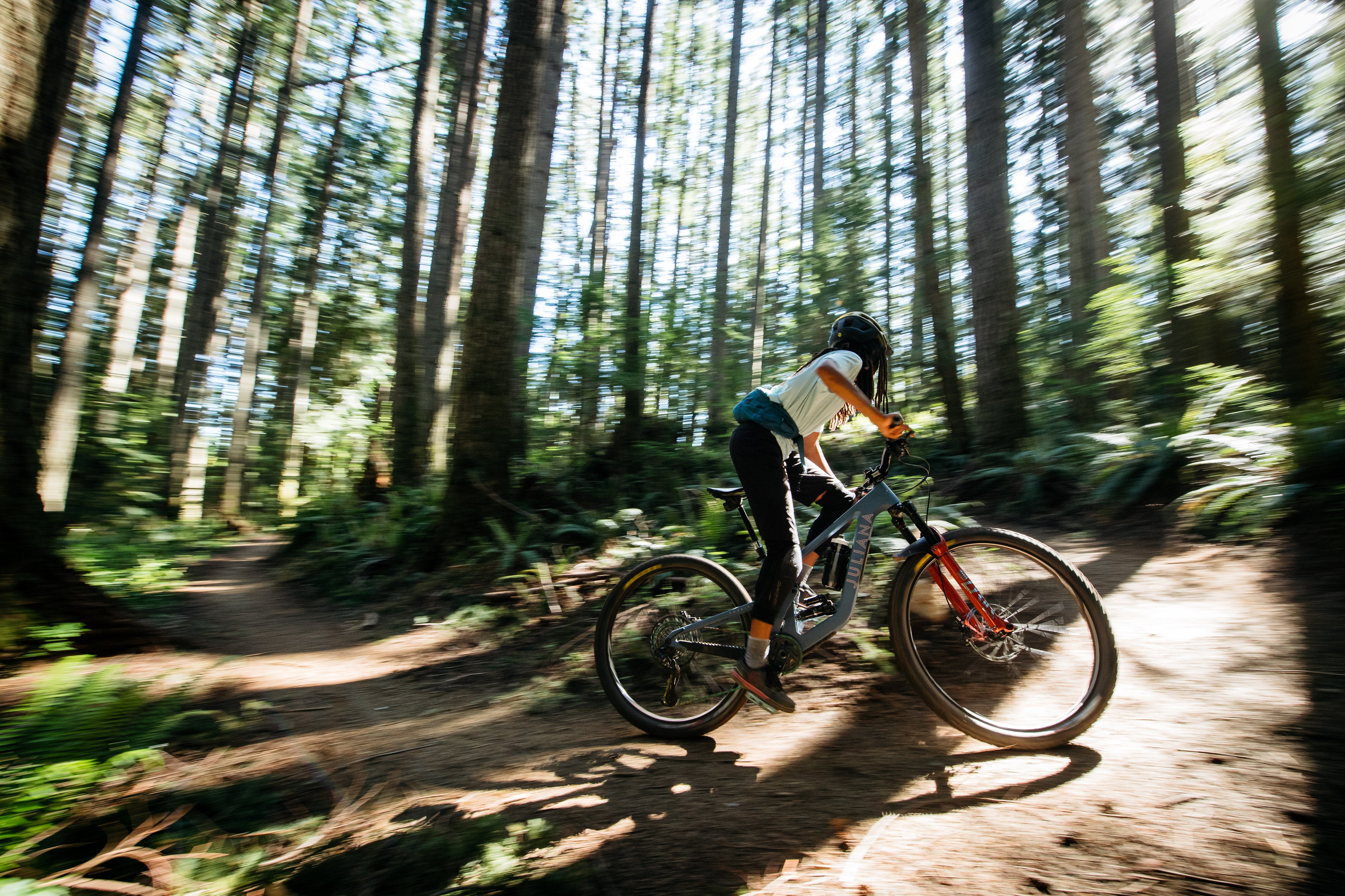 An action shot of a woman riding a Juliana Roubion mountain bike through a lush forest trail, navigating over roots and uneven ground, showcasing the bike’s agility and performance in natural terrain, available at Contender Bicycles.