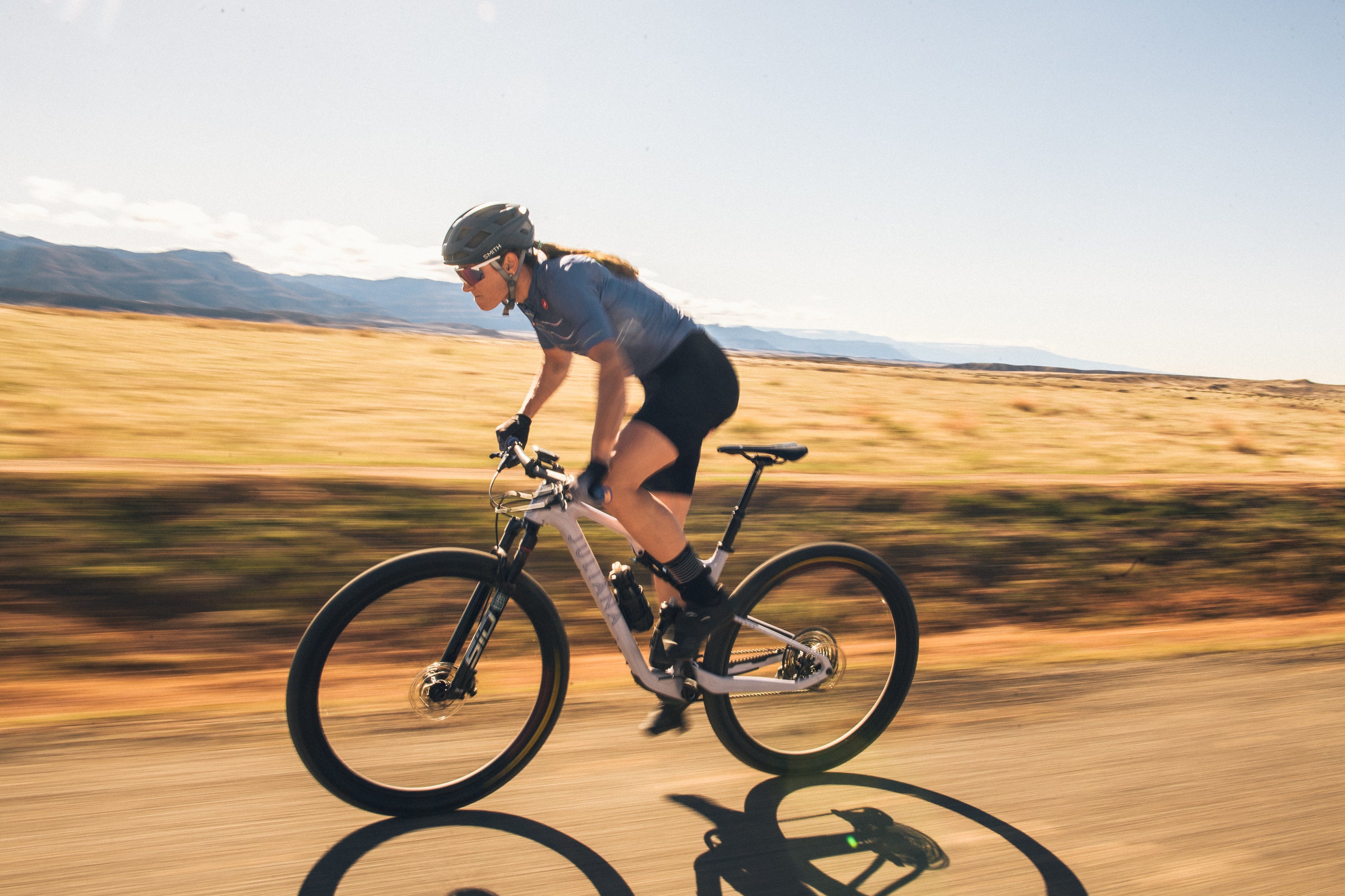 An action shot of a woman sprinting on a Juliana Wilder mountain bike through the high desert, with the bike’s wheels kicking up dust and majestic mountains in the background, highlighting the bike’s speed and performance on rugged terrain, available at Contender Bicycles.