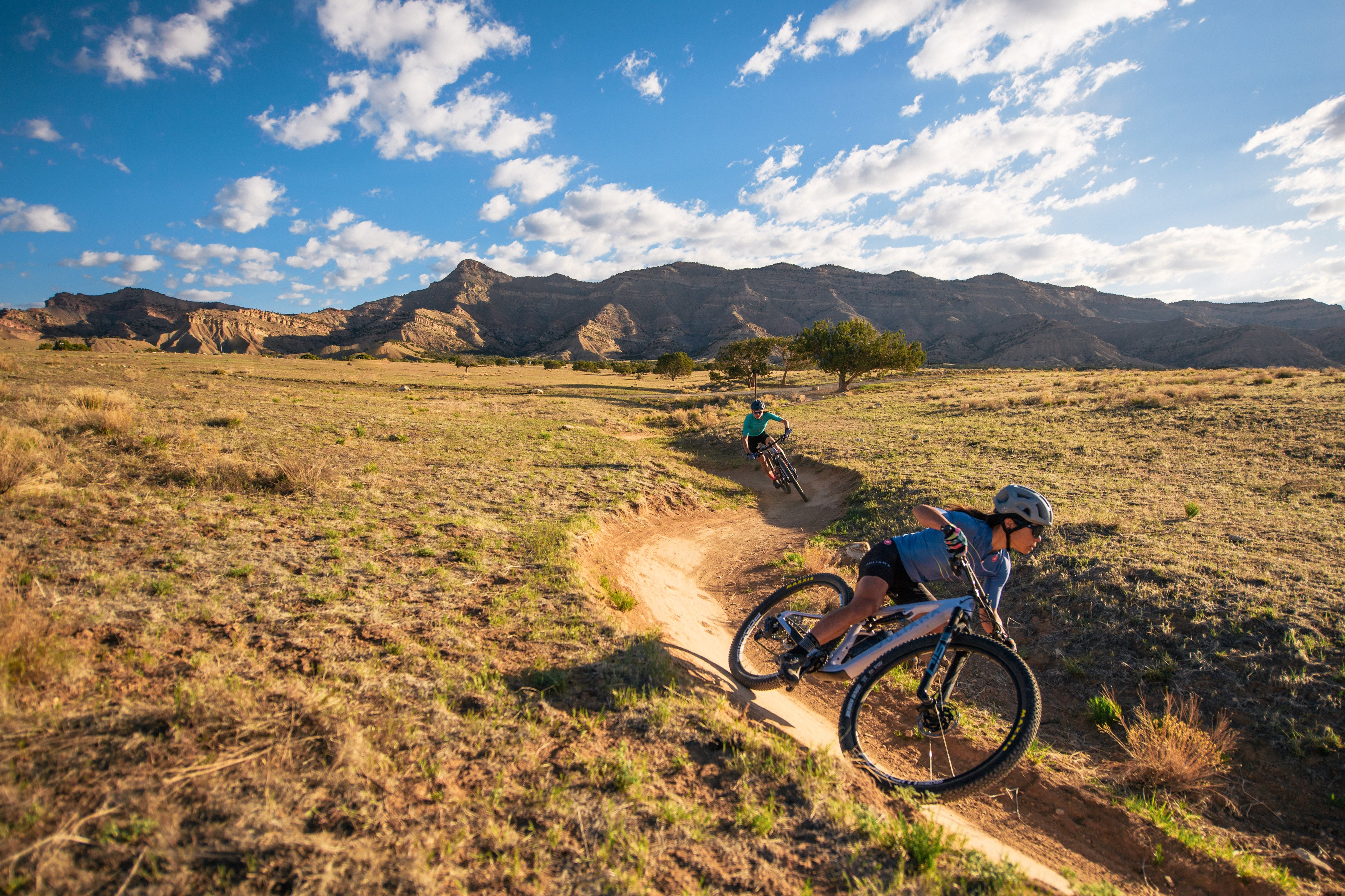 An action shot of two women riding Juliana mountain bikes on a trail, with one leaning into a sharp corner while the other follows, showcasing the bikes’ agility and performance on diverse terrain, available at Contender Bicycles.