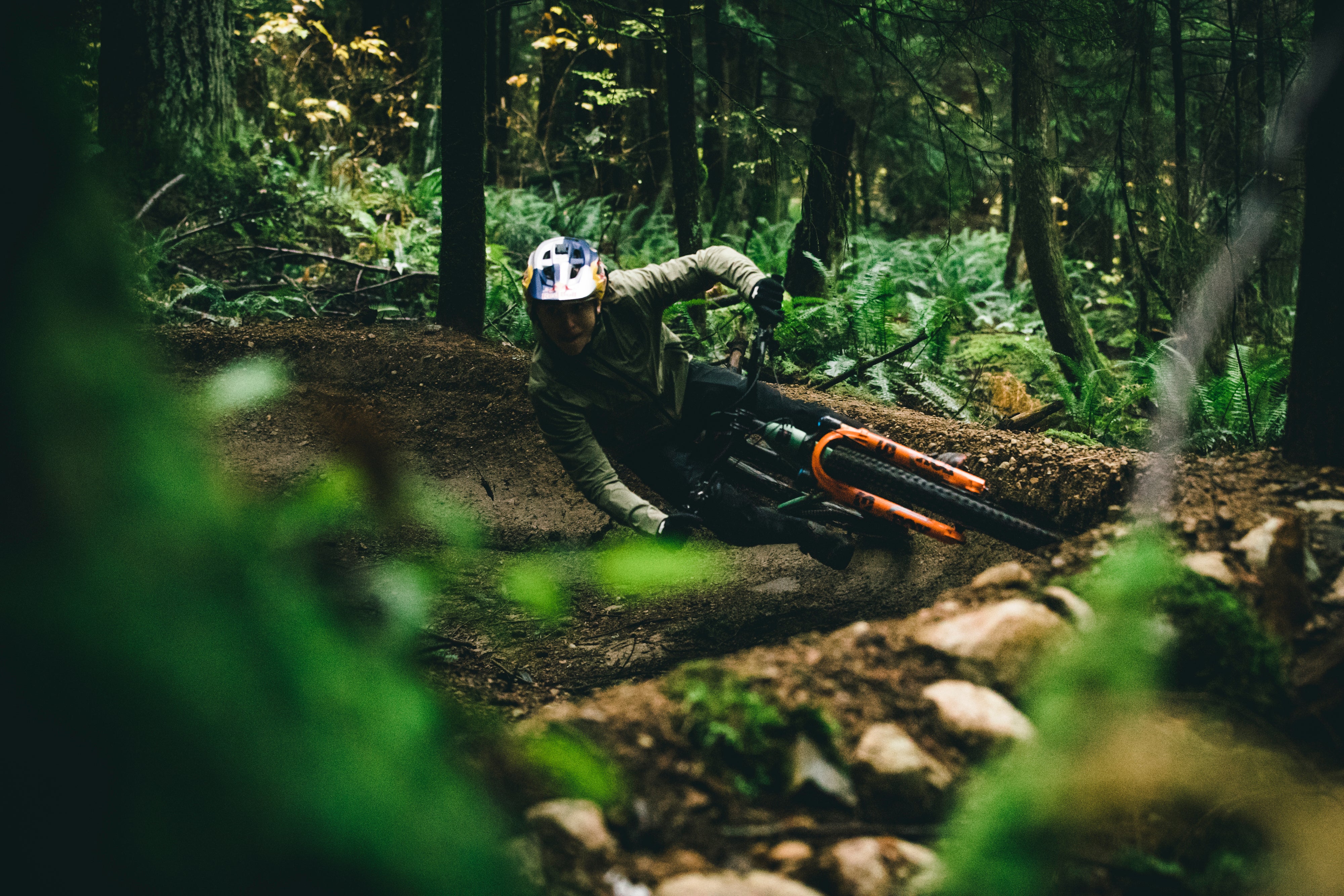 An action shot of a man carving a sharp corner on a Santa Cruz 5010 mountain bike, kicking up dirt on a scenic forest trail, surrounded by towering trees, available at Contender Bicycles.