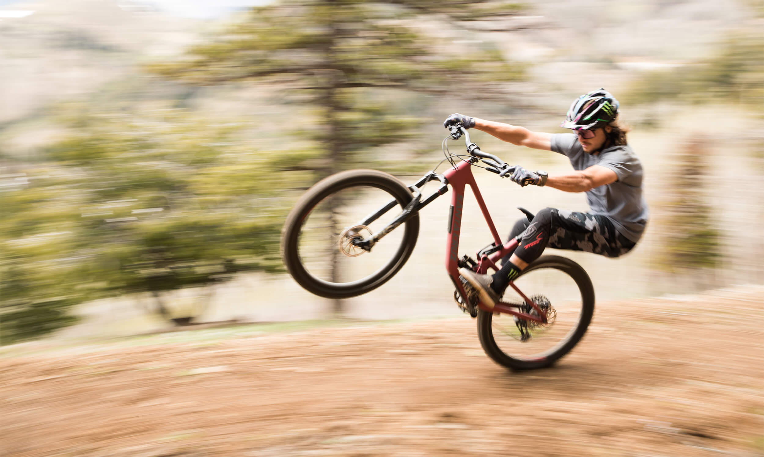 An action shot of a man performing a wheelie on a Santa Cruz 5010 mountain bike, surrounded by rugged desert terrain and cacti under a clear blue sky, available at Contender Bicycles.