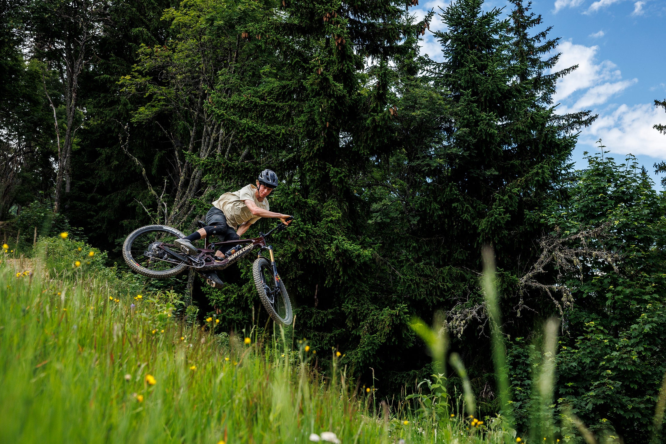 An action shot of a man launching off a jump on a Santa Cruz Bronson mountain bike, mid-air with a forest backdrop of tall trees and lush greenery, available at Contender Bicycles.