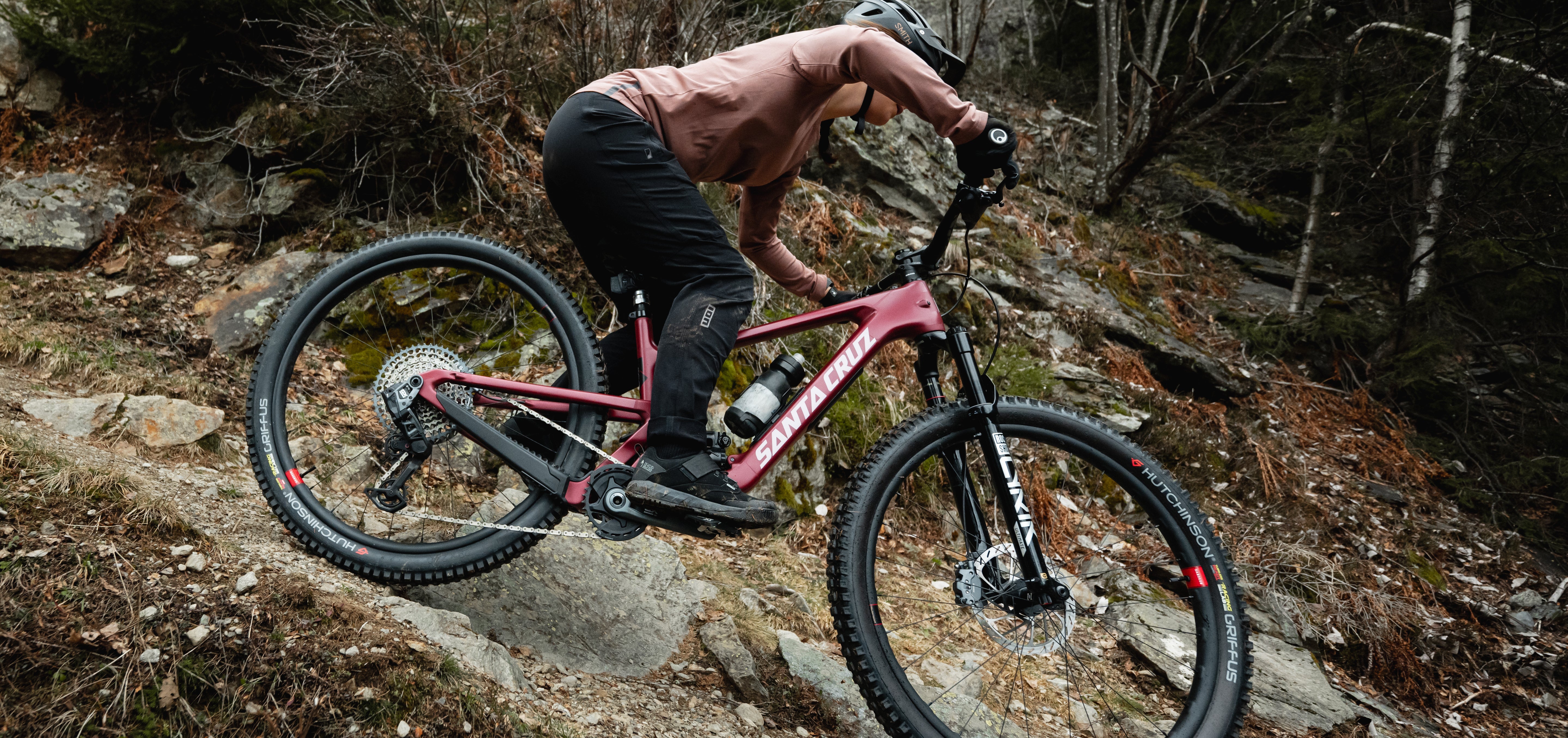 An action shot of a man riding a Santa Cruz Hightower down a rainy, rocky mountain trail, with water splashing and mist in the air, highlighting the bike's stability and performance in challenging conditions, available at Contender Bicycles.