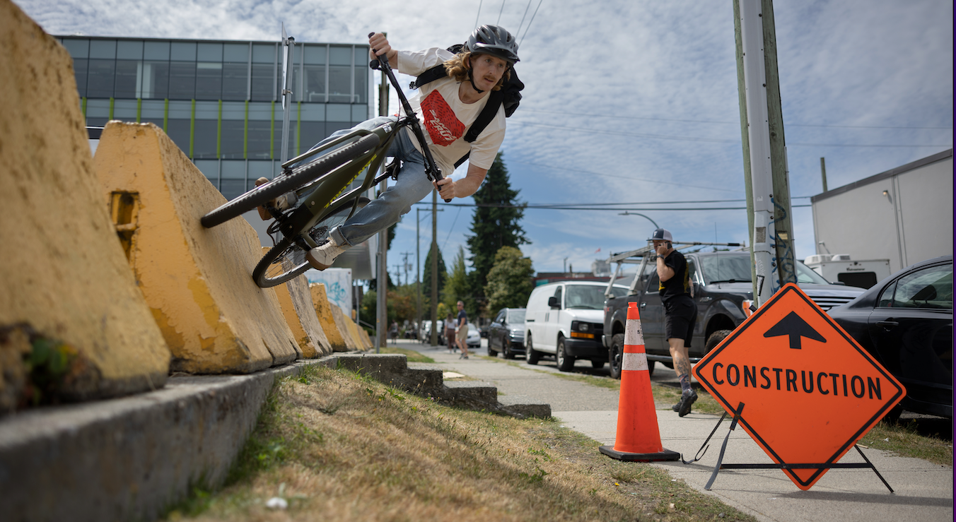 An action shot of a man launching off a jump on a Santa Cruz Stigmata electric bike, soaring through a construction site with scaffolding and machinery in the background, showcasing the bike’s power and agility in an urban landscape, available at Contender Bicycles.