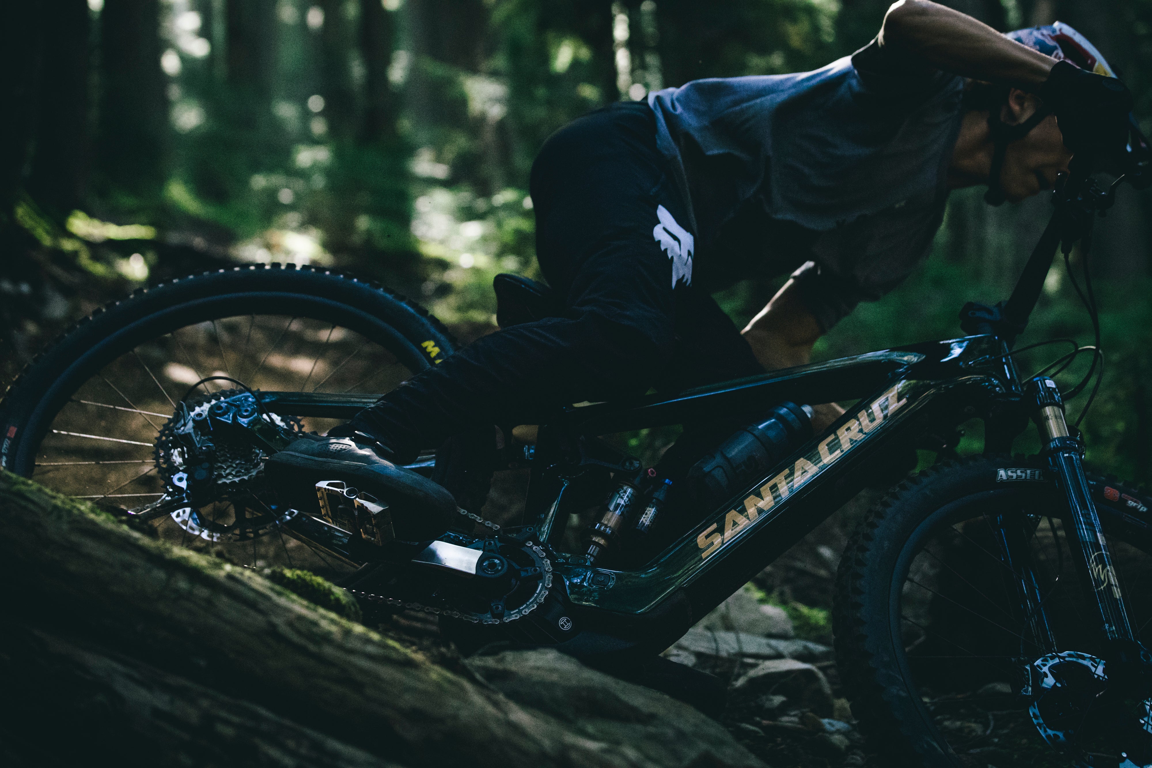 An action shot of a man riding a Santa Cruz Vala E-MTB through a forest trail, navigating loose rocks and rough terrain, showcasing the bike’s power and stability in challenging conditions, available at Contender Bicycles.