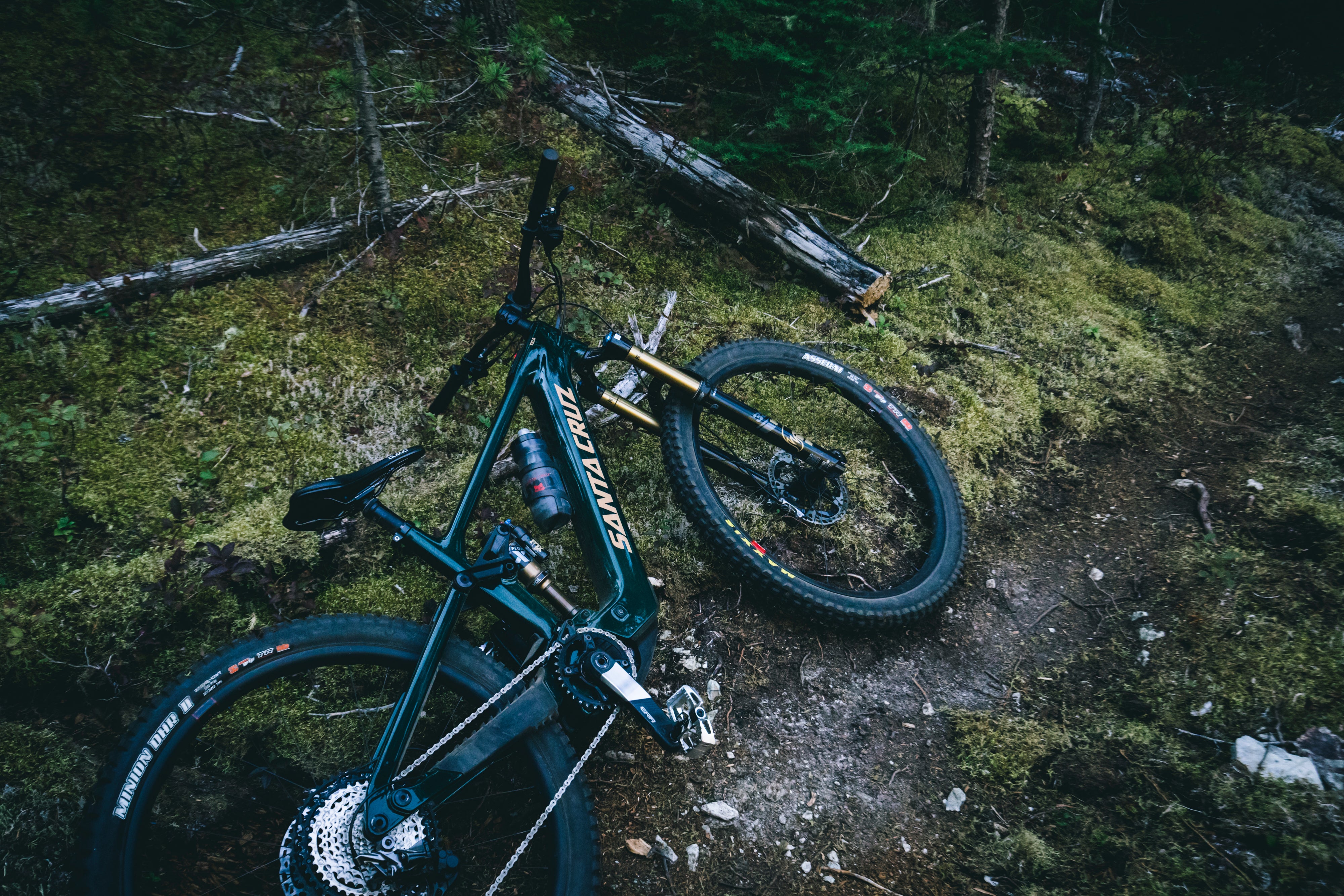 A still shot of a Santa Cruz Vala E-MTB laid on its side along a forest trail, surrounded by rough terrain and nature, highlighting the bike's rugged design, available at Contender Bicycles.