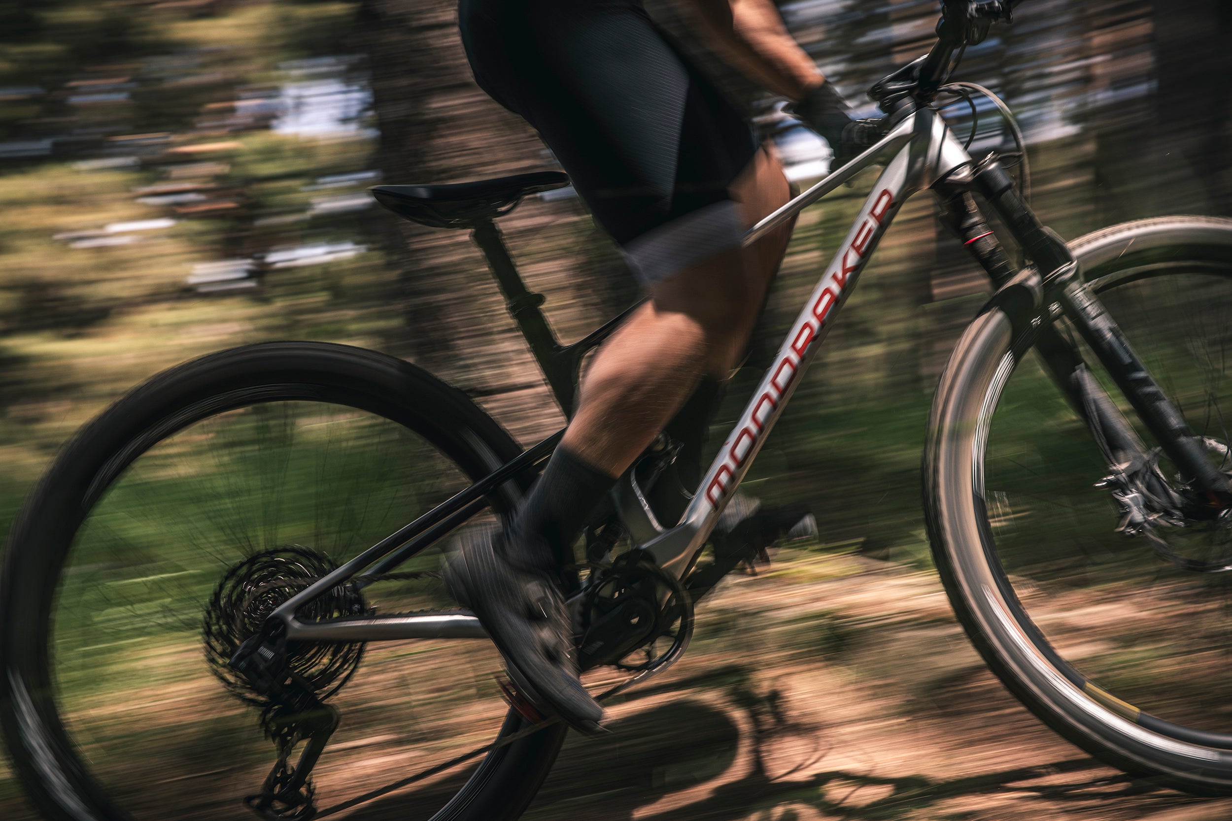 A man riding the black, gray, and red mondraker F-podium cross-country bike through a forest.