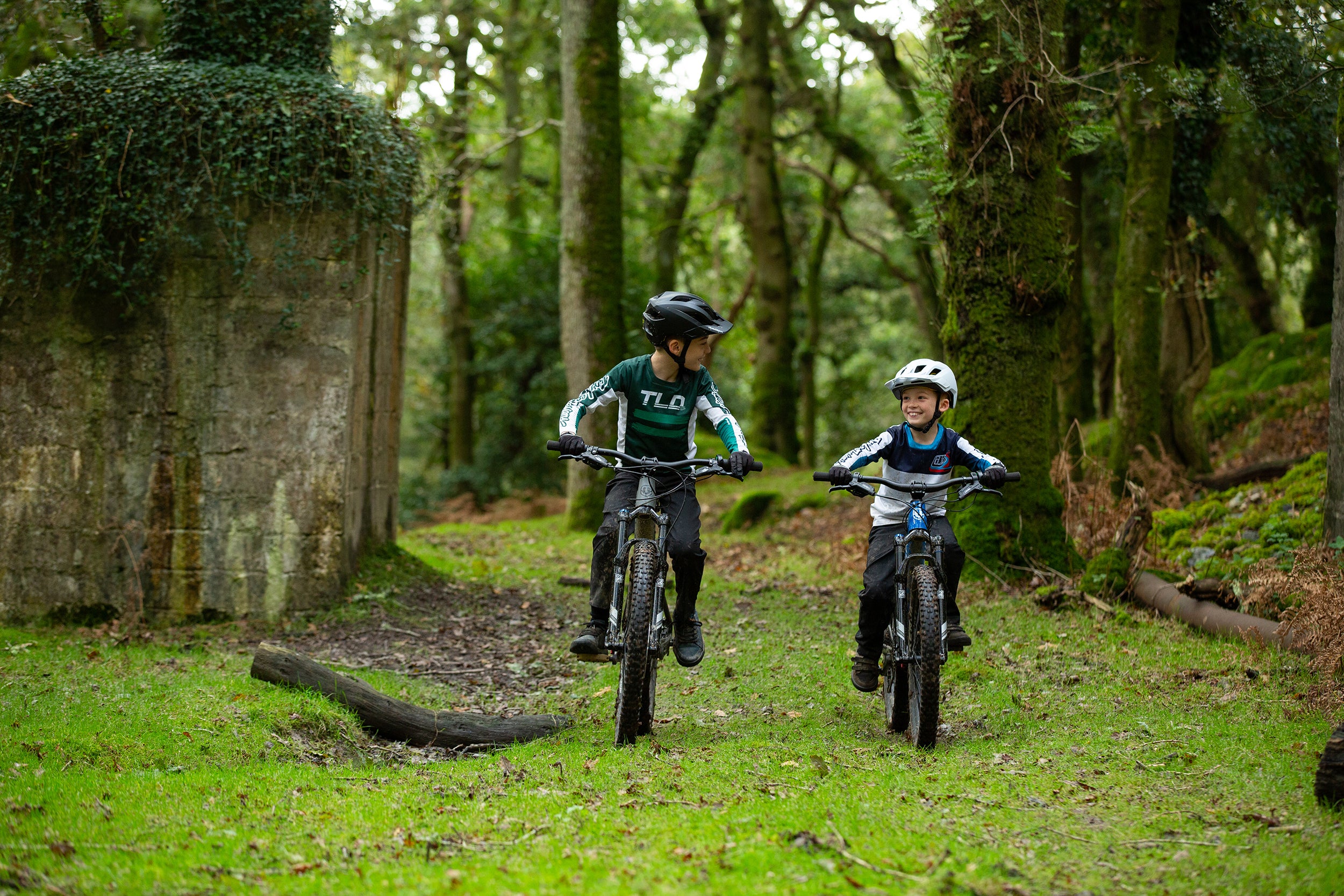 Two children riding the Mondraker F play, in a lush green forest, available at  Contender Bicycles.
