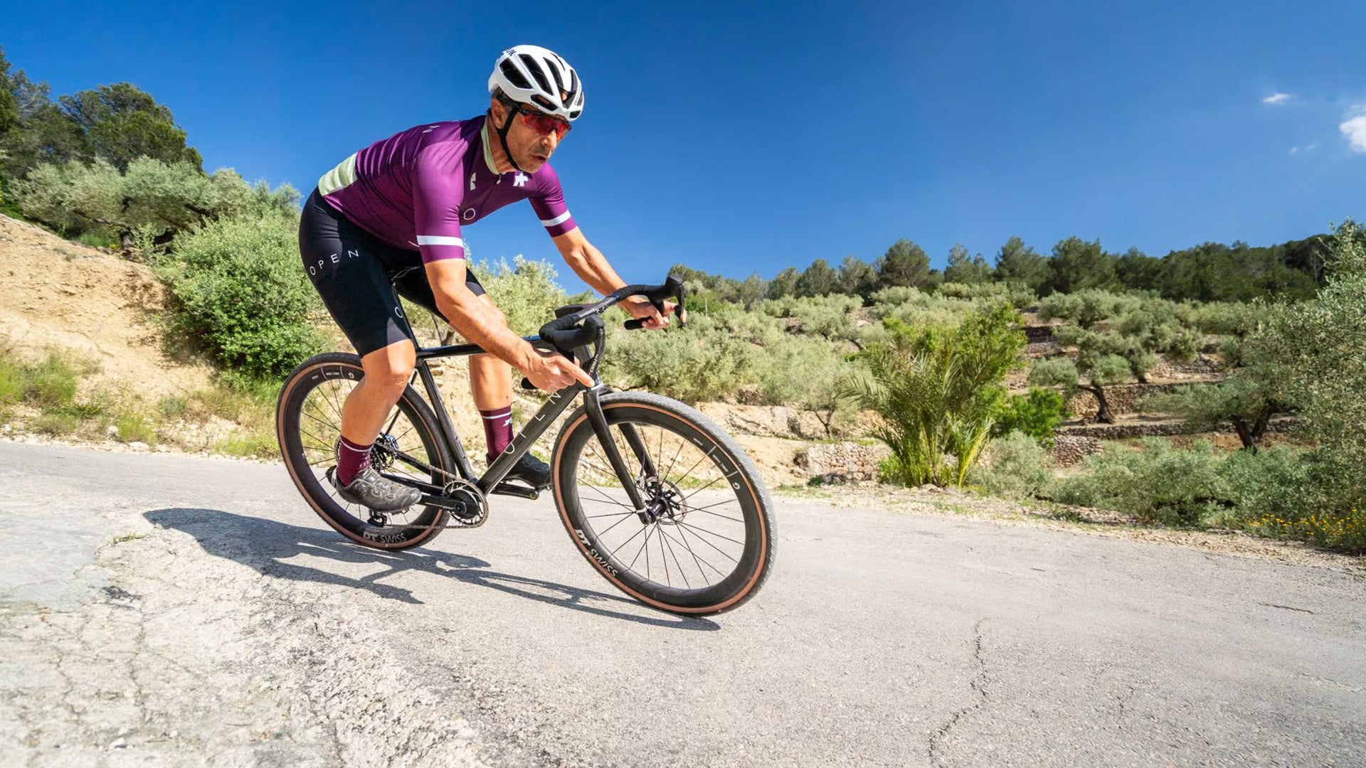 Cyclist in purple jersey and helmet riding a bicycle on a road with greenery in the background
