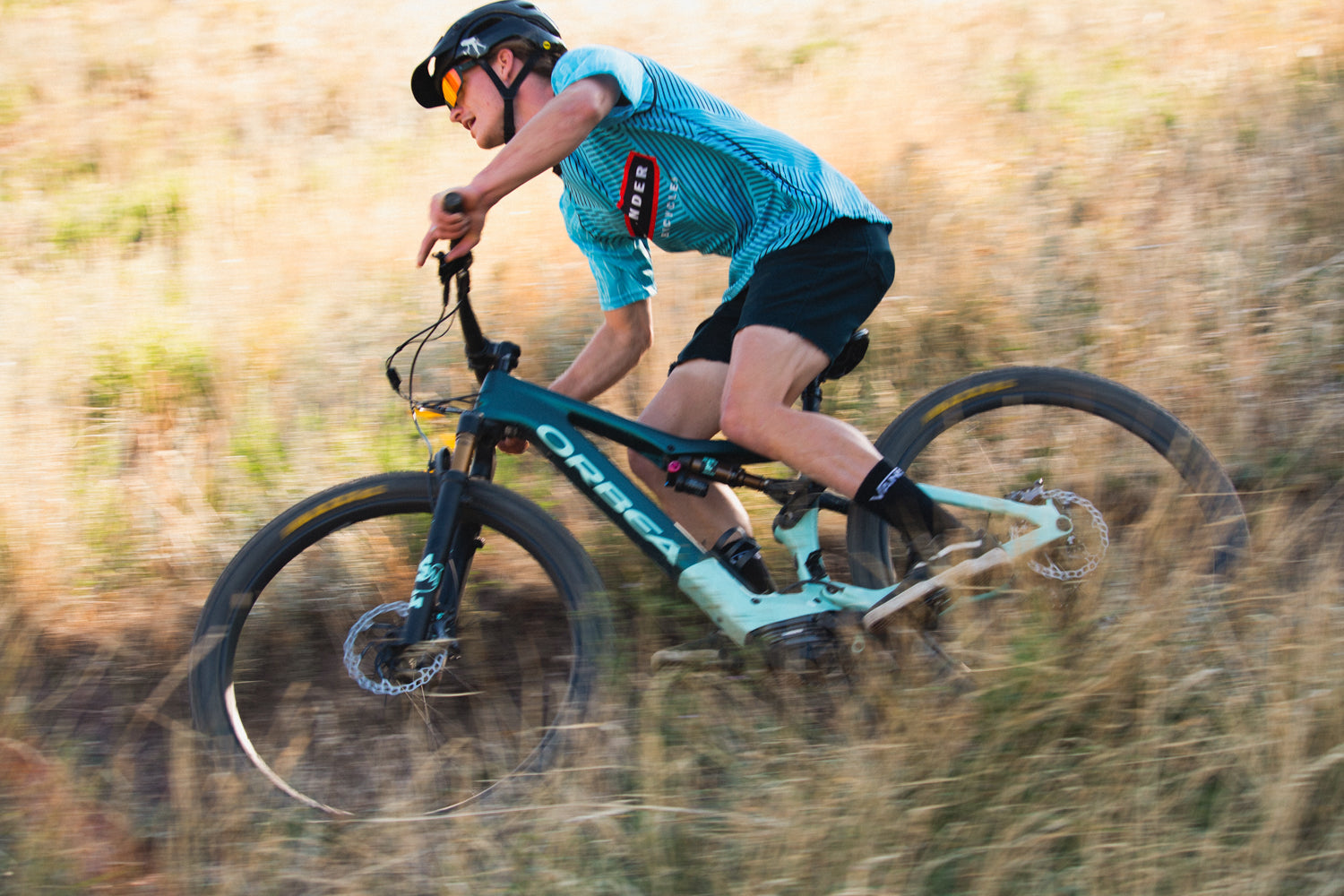 A man in a blue shirt riding the Orbea Rise E-mountain bike on a trail, surrounded by tall, brown grass. 