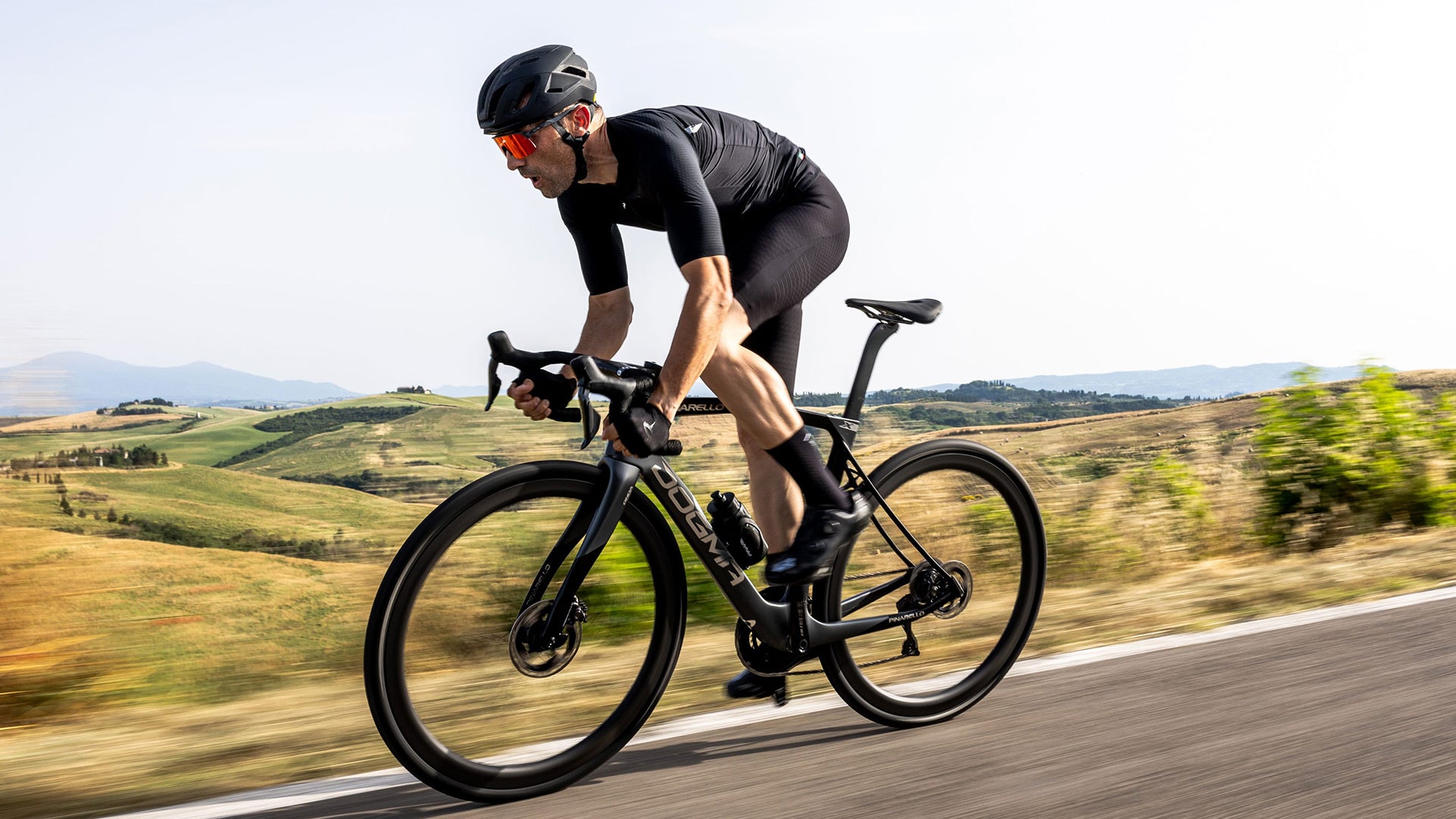 A man wearing all black riding a pinarello road bike against a countryside. 